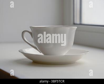 White ceramic cup and saucer on windowsill, light from window Stock Photo