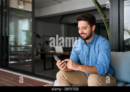 Happy smiling young business man holding smartphone sitting in office. Stock Photo