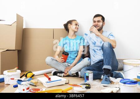 Happy smiling couple relaxing on floor with cup of coffee among construction tools and materials. House remodeling and interior renovation concept. Yo Stock Photo