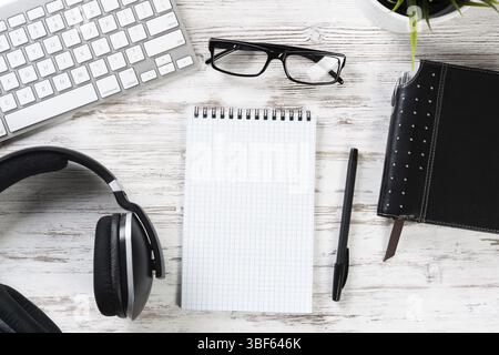 Top view modern office workspace. Flat lay white wooden desk with computer keyboard, headphones, glasses and opened blank spiral notepad with pen. Tim Stock Photo