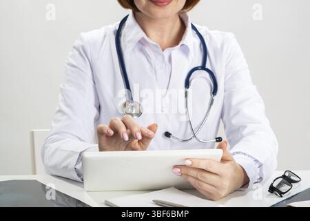 Close up female doctor hands using tablet computer. Family physician in white coat with stethoscope sitting at desk. Professional medical diagnosis an Stock Photo