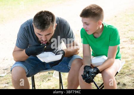 A man and boy sit on chairs outside, wearing black gloves while eating from disposable containers. They appear to be enjoying a casual meal. Stock Photo