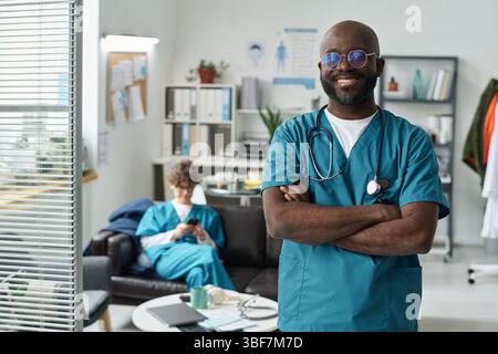 African American doctor standing with arms crossed, smiling confidently in modern medical office, while a nurse sits on couch in background, busy with paperwork Stock Photo