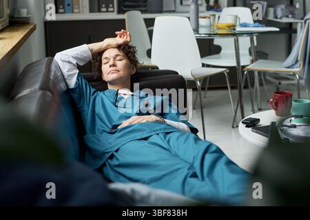 Exhausted healthcare worker resting on couch during break, person reclined with arm over head in break room. Medical uniform and equipment visible in background Stock Photo