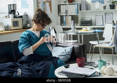 Nurse in scrubs sitting in a break room, analyzing patient records in a hospital setting with various medical tools and paperwork. Visible cups and gadgets on table create a lived-in environment Stock Photo