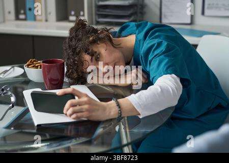 Exhausted healthcare worker wearing scrubs resting head on desk during break in a medical office environment. Coffee mug and stack of paperwork visible on the table Stock Photo