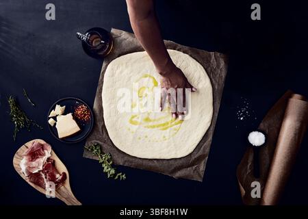 Hand spreading olive oil on pizza dough surrounded by toppings Stock Photo