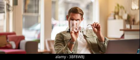 Portrait of man with new anti blue light glasses, looking through the lense at his computer screen, sitting in cafe with laptop and testing eyewear Stock Photo