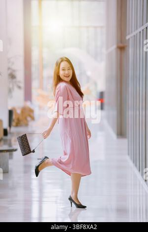 Cheerful Southeast Asian woman in pink dress smiling and posing playfully with one leg up in bright indoor hallway with bokeh background Stock Photo