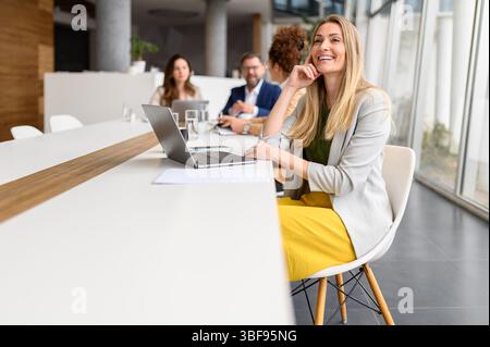 Smiling female manager working online over laptop while business colleagues discussing in the background Stock Photo