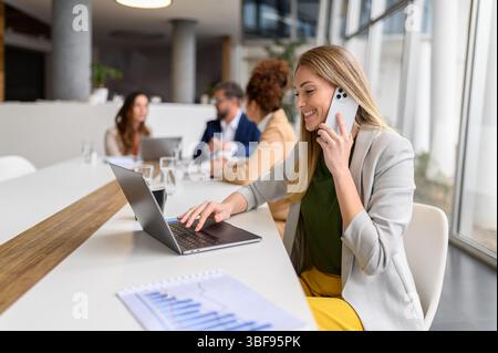 Smiling female entrepreneur working on laptop and discussing project details over call in meeting with professionals collaborating in the background Stock Photo