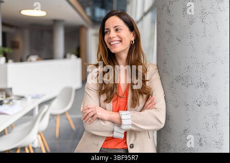 Portrait of confident businesswoman with arms crossed smiling and looking away while standing by column in office Stock Photo