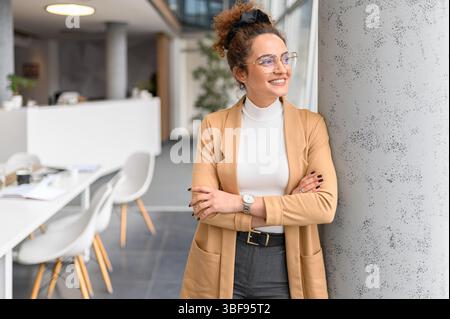 Female entrepreneur with arms crossed looking away and thinking business ideas while leaning on column in office Stock Photo
