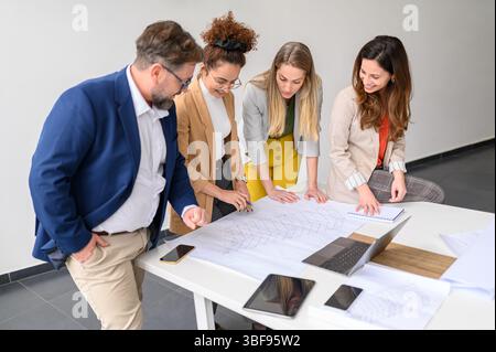 Smiling businesswoman explaining blueprint and planning project with colleagues at table in meeting room Stock Photo