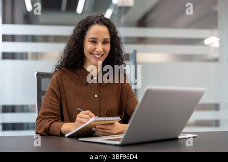 A woman with curly hair smiles while writing notes during an online meeting in the office. Her laptop sits open in front of her. Stock Photo