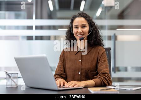 A smiling customer service representative wearing a headset in a modern office setting, using a laptop at a desk. Stock Photo