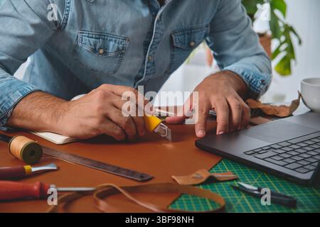 Shoemaker cutting leather with cutter next to laptop in his workshop Stock Photo