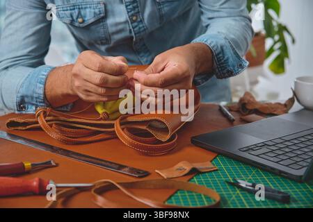 Leather craftsman carefully stitching a handmade bag in his workshop, surrounded by tools and technology Stock Photo