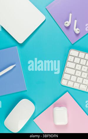 Top view of white keyboard, mouse, touchpad, earphones case, pen on blue background. Purple, pink and blue notebooks, modern office, school. Stock Photo