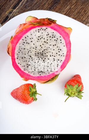 Close up of white plate with half of dragon fruit with black seeds and two fresh strawberries on wooden background. Stock Photo