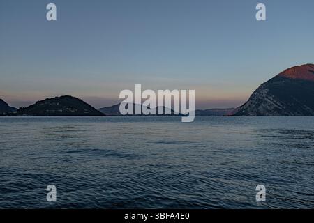 The first light of dawn paints the sky in warm colours, reflecting on the mountains surrounding the calm waters of Lake Iseo, in Lombardy, Italy. Stock Photo