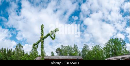 Traditional midsummer celebration featuring a maypole adorned with flowers and leaves, rising against a cloudy sky in Gammlia, Umea, Sweden, amidst lu Stock Photo
