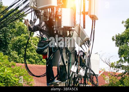 A technician performs maintenance on a communication tower, managing cables and equipment while ensuring safety during the height work in an urban set Stock Photo