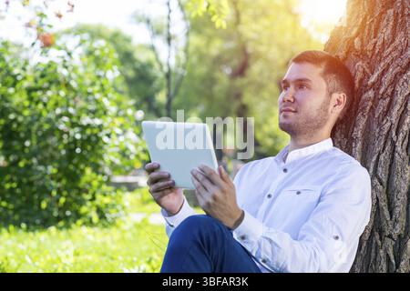 Dreamy man using tablet computer under tree in park on sunny day. Handsome businessman looking over tablet screen. Dreamy man sitting on green grass l Stock Photo