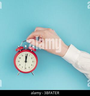 Creative flat lay concept top view of woman hand in white shirt holding the red vintage alarm clock on bright blue turquoise square color paper backgr Stock Photo