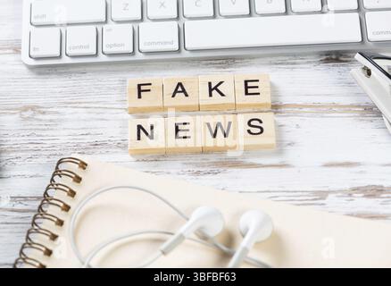 Fake news concept with letters on cubes. Flat lay vintage wooden desk with computer keyboard, headphones, and notebook. Disinformation media technolog Stock Photo