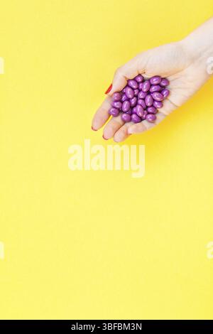 Woman hand holding purple pills handful isolated on bold yellow background with copy space. Template for feminine beauty blog social media. Female hea Stock Photo