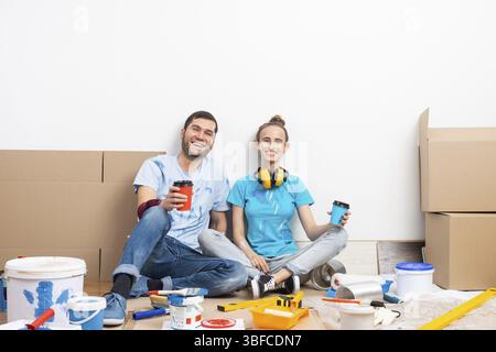 Happy smiling couple relaxing on floor with cup of coffee among construction tools and materials. House remodeling and interior renovation concept. Yo Stock Photo
