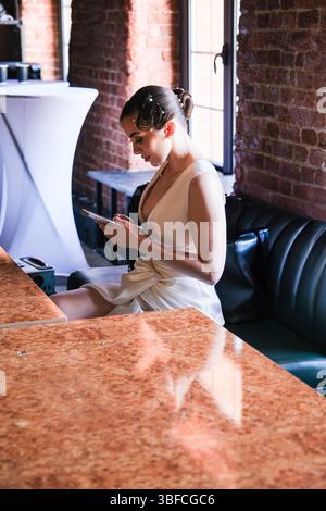 moscow, russia, 22.05.2025 A woman in a white dress is sitting at a table with a tablet in front of her. She is reading or writing something on the ta Stock Photo