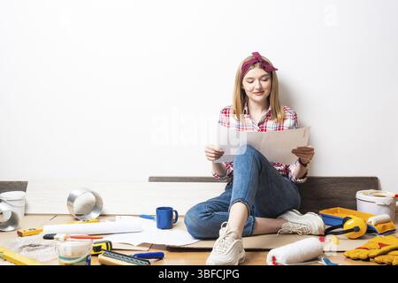 Smiling girl sitting on floor and holding paper blueprint. Home remodeling and house interior redesign. Construction tools and materials for building. Stock Photo