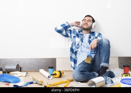 Handsome bearded man listening to music in headphones at home. Home remodeling after moving concept. Painting tools and material on floor. Young cauca Stock Photo