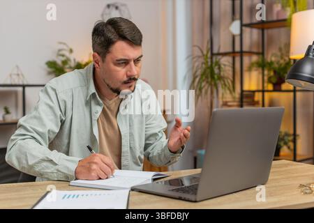 Young man writing down or taking down notes while attending online video class lessons or office meeting on laptop computer at home office desk. Guy freelancer support services looking at webcam. Stock Photo