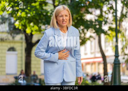 Sad mature businesswoman boss in formal suit suffering from stomach pain while walking on city town street. Blonde middle-aged woman employer has poisoning, spasms, menstruation or digestive problems Stock Photo