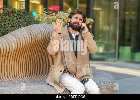 Indian young businessman tourist having bad stressful news calling on smartphone. Sad frustrated confused Hispanic bearded guy have problem unpleasant remote phone conversation on downtown city street Stock Photo