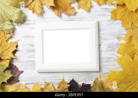 Blank rectangular photo frame lies on vintage wooden desk with bright autumn foliage. Flat lay with autumn leaves on white wooden surface. Simple whit Stock Photo