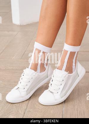 White sneakers paired with transparent socks featuring two vertical stripes, worn by a person standing on wooden flooring, showcasing a casual and Stock Photo