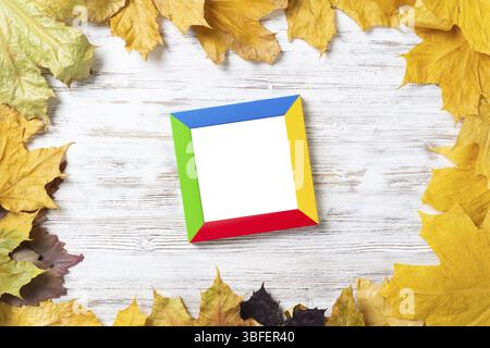 Blank rectangular photo frame lies on vintage wooden desk with bright autumn foliage. Flat lay with autumn leaves on white wooden surface. Simple colo Stock Photo
