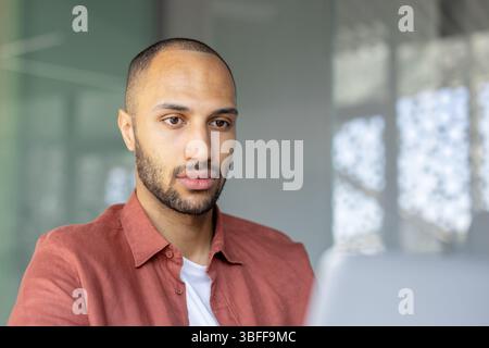 A focused, young man with a beard looks at a laptop screen, in a workspace with natural light filtering through the background. Stock Photo