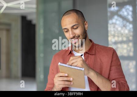 A young man is seen writing in a notepad with a pen, looking focused and content, probably at work. Stock Photo