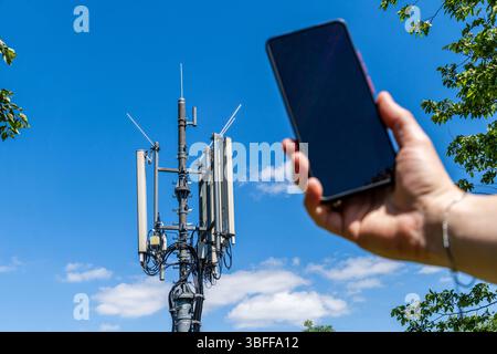 Munich, Bavaria, Germany - May 30, 2025: A man holds a smartphone in the direction of a mobile phone mast, symbolic image for network coverage, reception quality, connection speed and the importance of modern communication technology *** Ein Mann hält ein Smartphone in Richtung eines Mobilfunk-Sendemasts, Symbolbild für Netzabdeckung, Empfangsqualität, Verbindungsgeschwindigkeit und die Bedeutung moderner Kommunikationstechnik Stock Photo