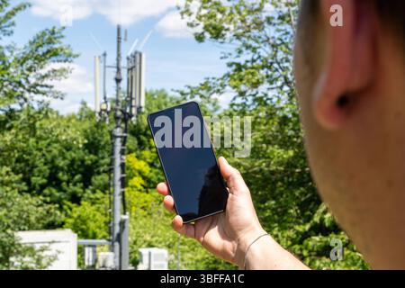 Munich, Bavaria, Germany - May 30, 2025: A man holds a smartphone in the direction of a mobile phone mast, symbolic image for network coverage, reception quality, connection speed and the importance of modern communication technology *** Ein Mann hält ein Smartphone in Richtung eines Mobilfunk-Sendemasts, Symbolbild für Netzabdeckung, Empfangsqualität, Verbindungsgeschwindigkeit und die Bedeutung moderner Kommunikationstechnik Stock Photo