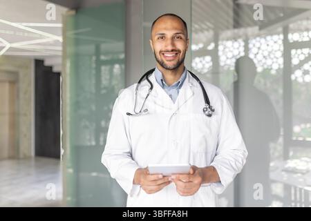 A smiling male doctor in a white coat, holding a tablet, standing in a modern healthcare setting. He looks friendly and approachable, ready to help. Stock Photo