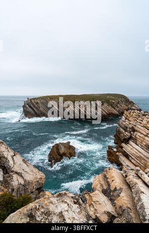 A unique isolated island with prominent layered rock formations is visible in the ocean, with waves breaking around its base and foreground cliffs Stock Photo