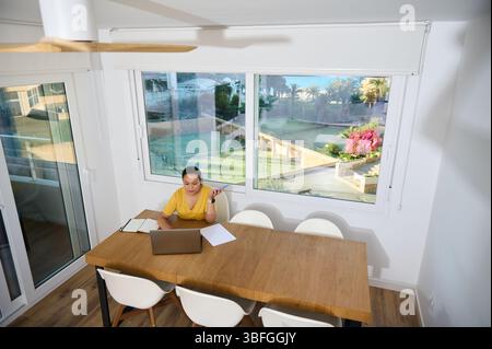 A businesswoman in a modern home office works on a laptop against a backdrop of lush gardens seen through large windows, creating a serene and product Stock Photo