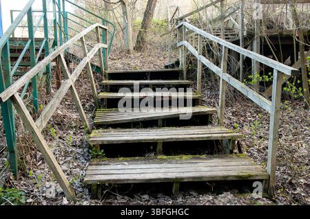A decaying wooden staircase seen from below, with patches of green moss on some steps. Beside it, a rusty metal staircase with traces of faded green p Stock Photo