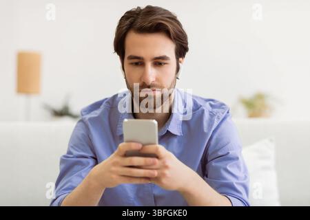 Serious Man Using Smartphone Wearing Earbuds Sitting On Sofa Indoors Stock Photo
